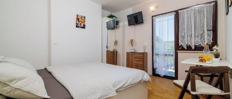 Un dormitorio con cama, escritorio con silla, televisión montada en la pared y ventana con cortinas de encaje.