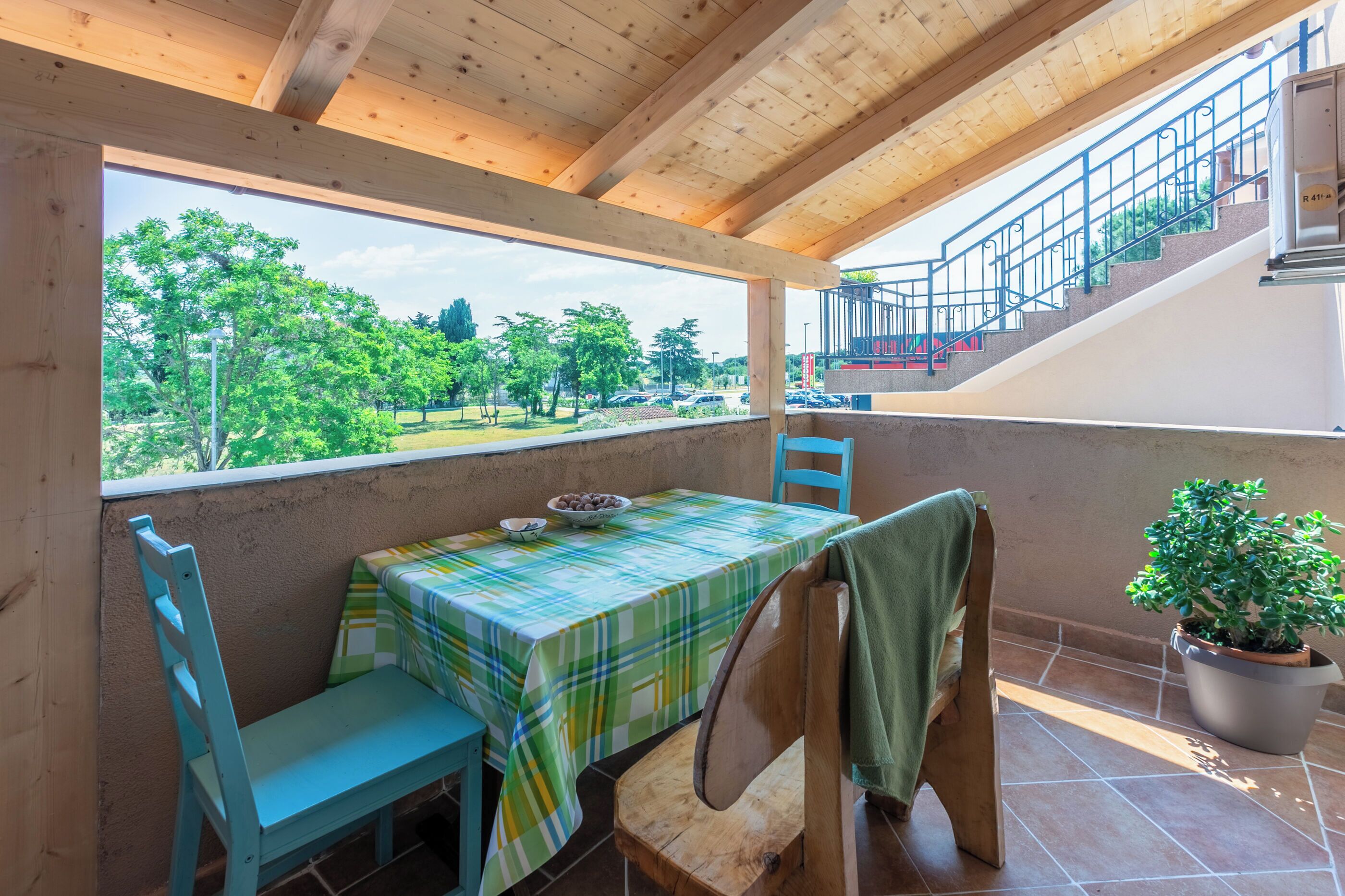Een balkon met een tafel en stoelen, een plant in pot en uitzicht op groen en een parkeerplaats.