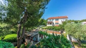 A two-story building with a red-tiled roof, a balcony on the upper floor, and a garden with various plants and a tree. - Apartments Armando (Fazana)