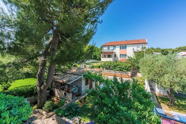 A two-story building with a red-tiled roof, a balcony on the upper floor, and a garden with various plants and a tree.