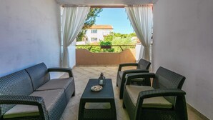 A balcony with wicker furniture, a table with a lantern, and a view of a house and greenery. - Apartments Armando (Fazana)