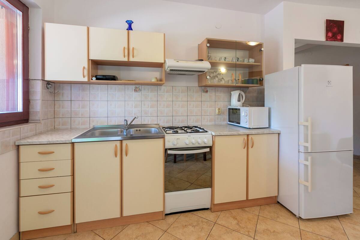 A kitchen with beige cabinets, a stove, a microwave, a refrigerator, and a window with blinds.