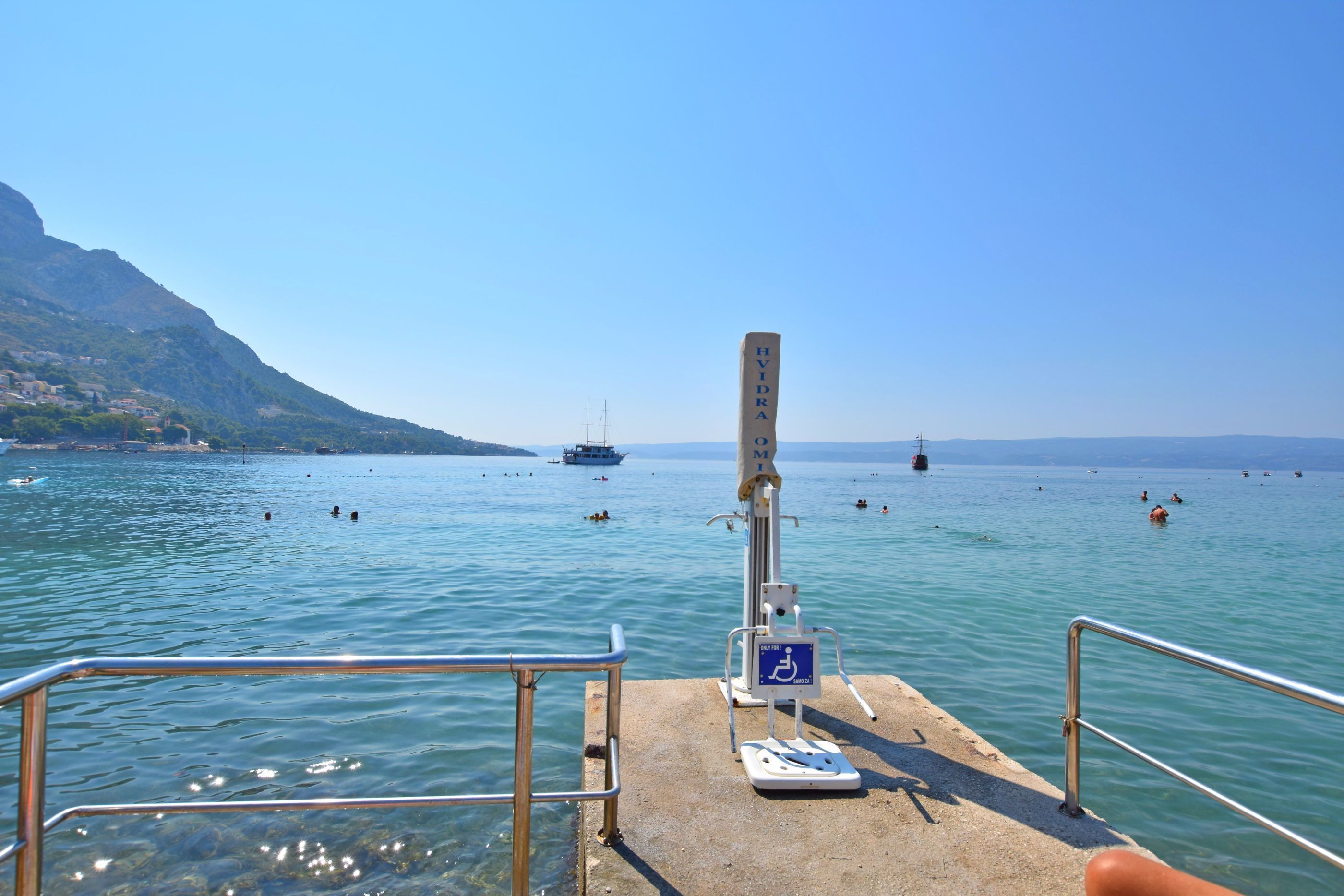 Un muelle con un puesto de salvavidas, un cartel de accesibilidad para sillas de ruedas, y una vista despejada del mar con barcos y personas nadando.