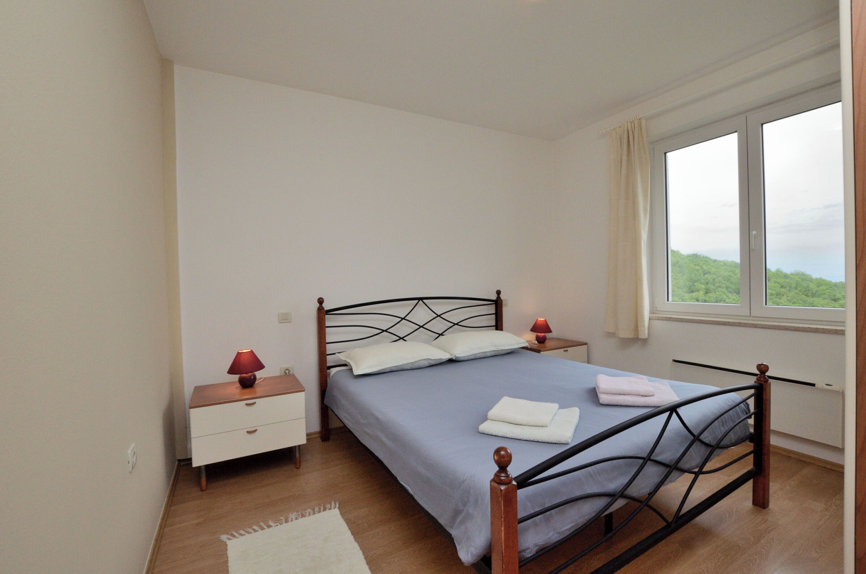 Ein Schlafzimmer mit Bett, Nachttisch, Lampe, Fenster mit Blick auf Grünflächen und einer weißen Wand.