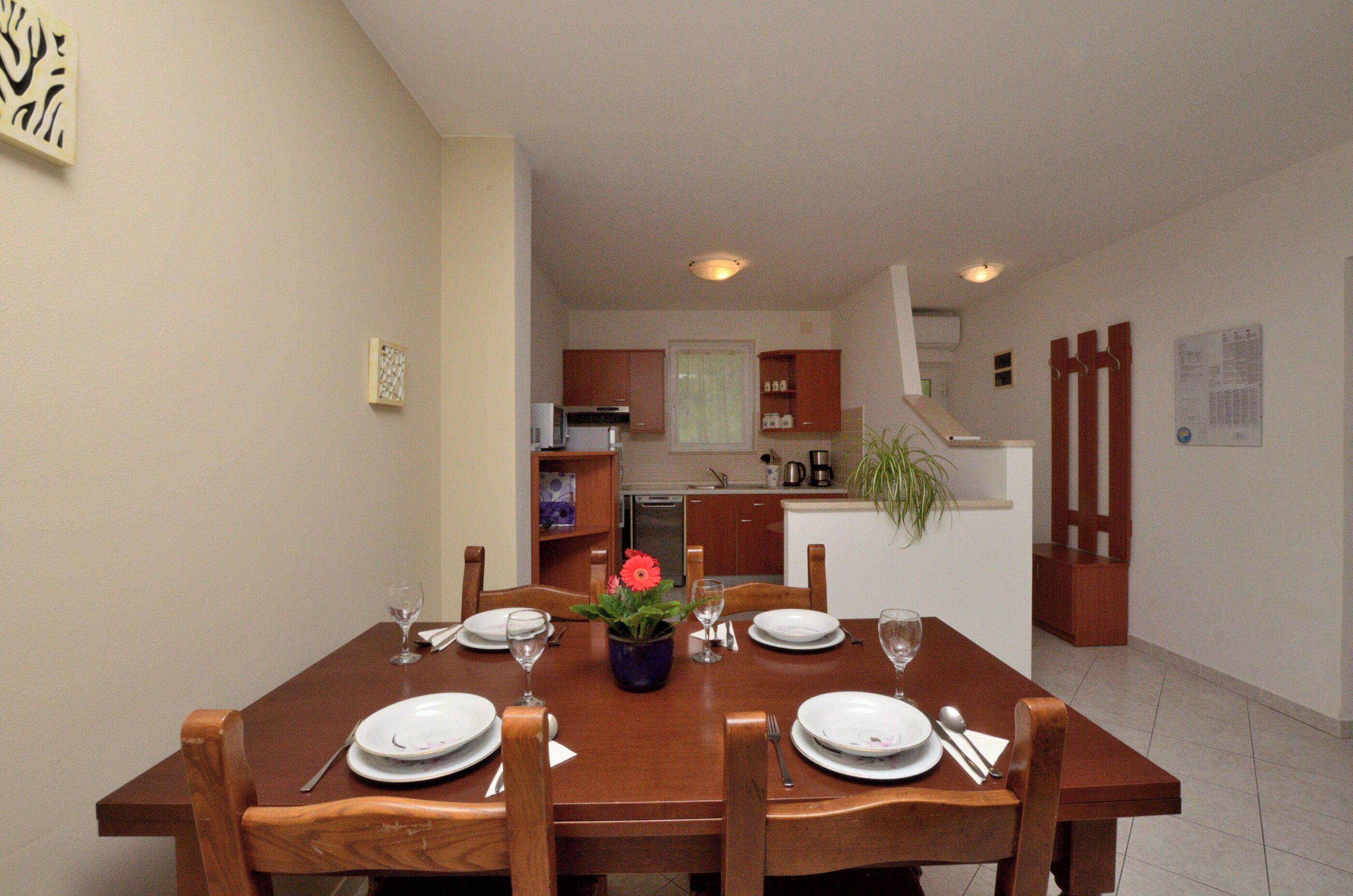 A dining area with a wooden table set for four, a kitchen in the background, and a staircase leading to another level.