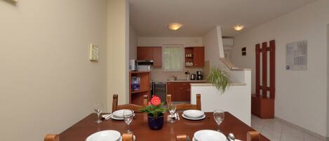 A dining area with a wooden table set for four, a kitchen in the background, and a staircase leading to another level.