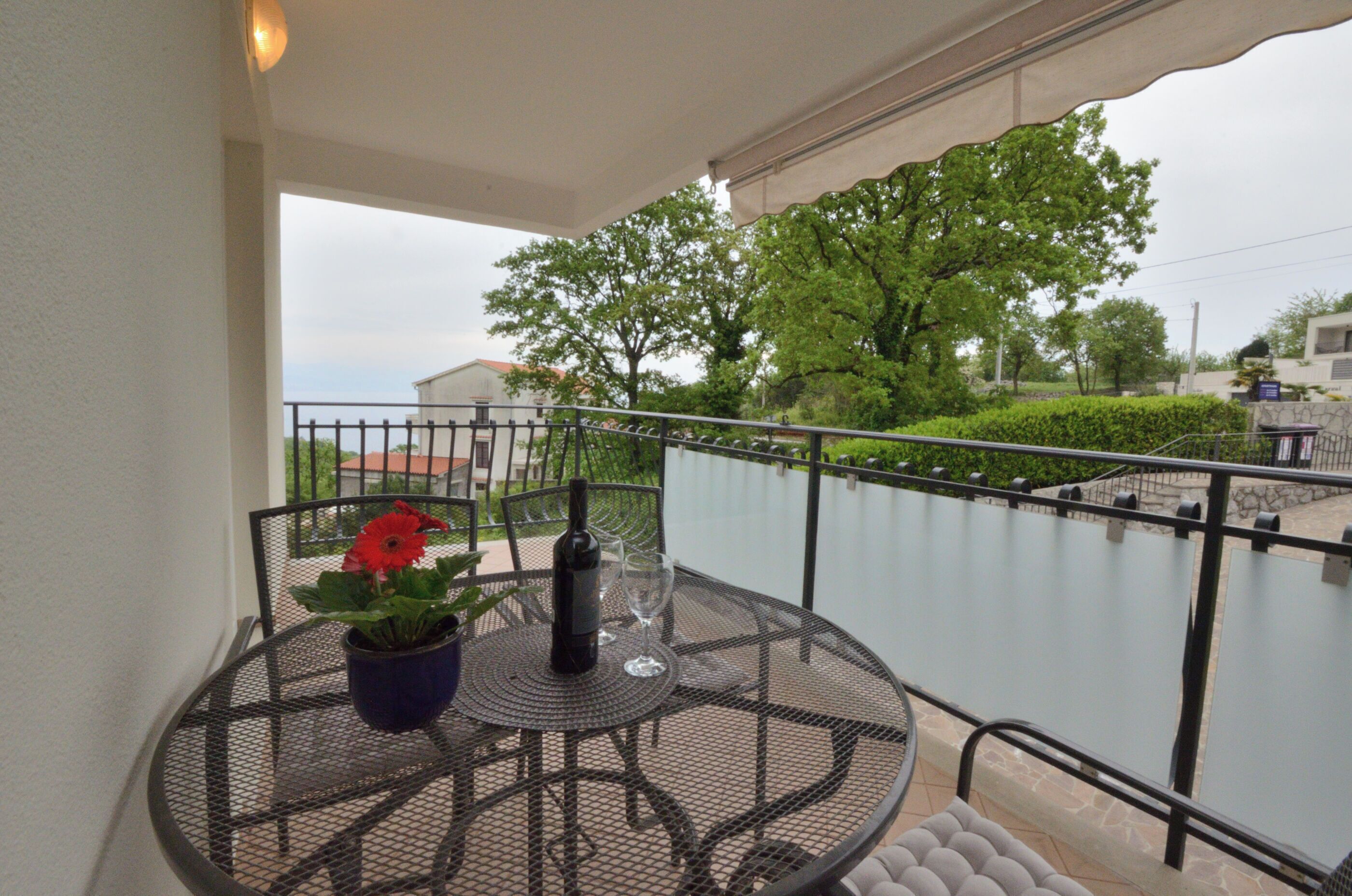 A round metal table with a potted plant and a bottle, set on a balcony with a glass railing overlooking a garden and a building.
