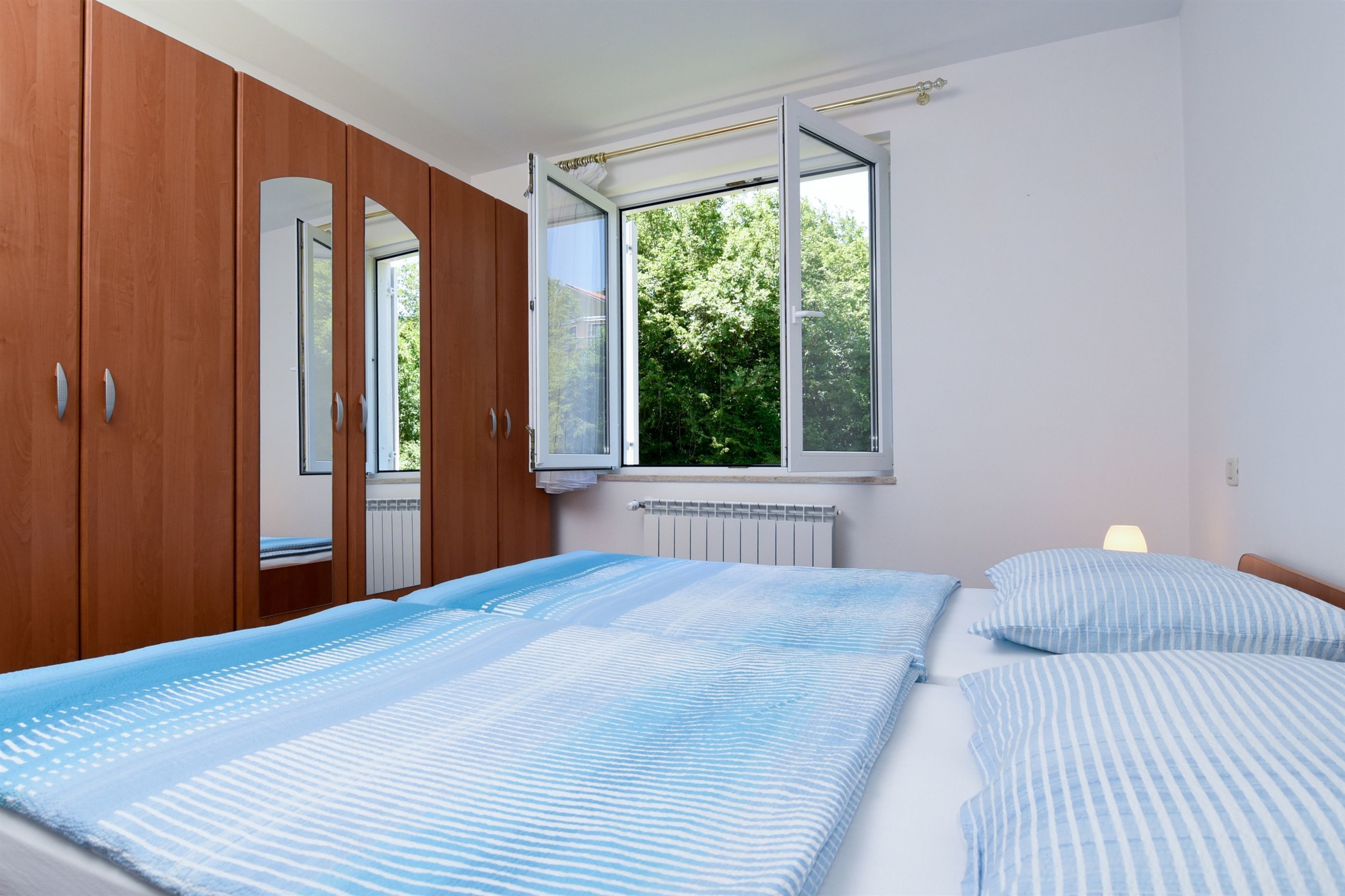 A bedroom with two beds, wooden wardrobe, and a window with a view of greenery.