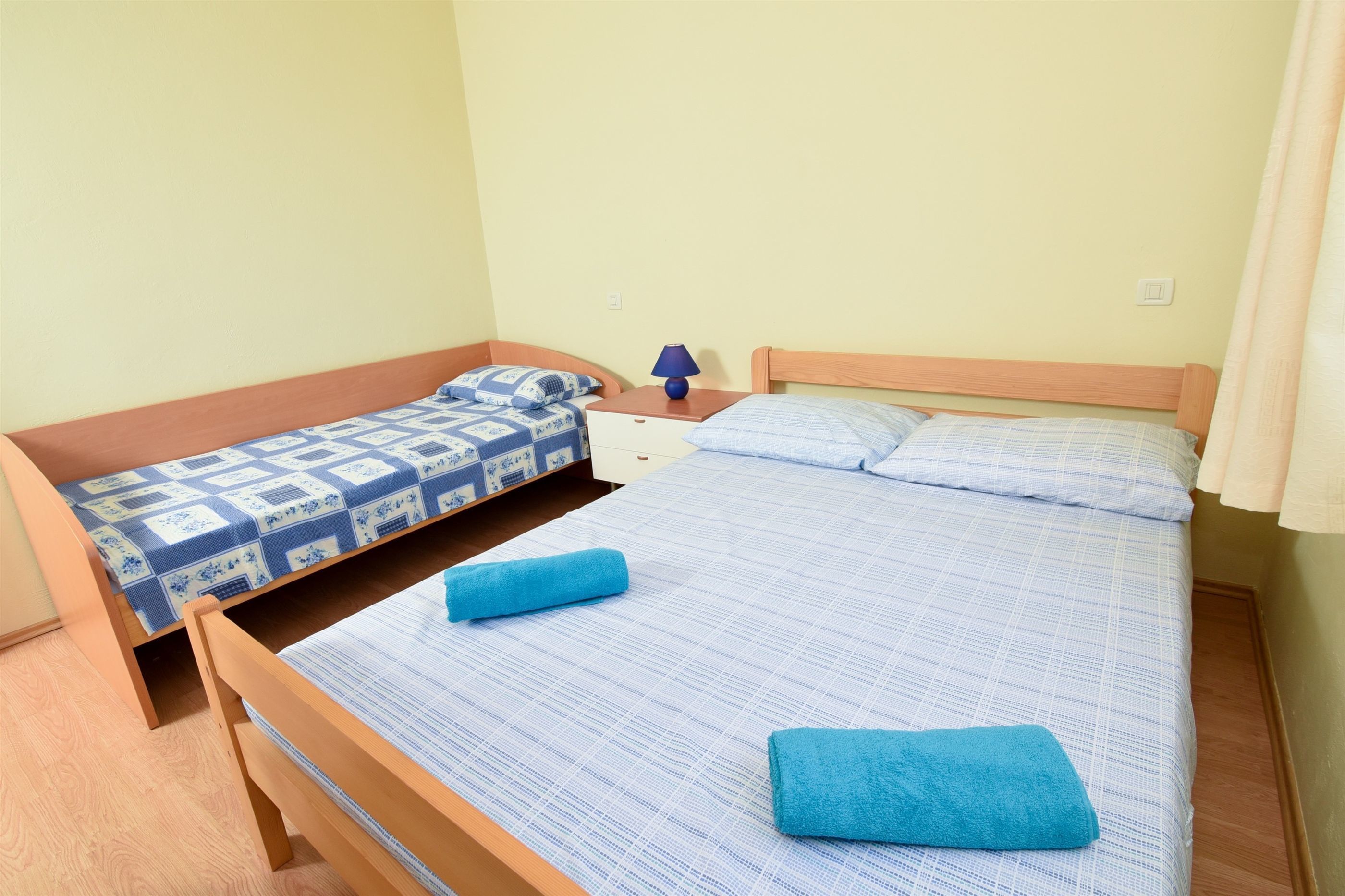 Ein Zimmer mit zwei Betten, eines mit einem karierten blau-weißen Überwurf und das andere mit einem schlichten blauen Überwurf. Dazwischen steht ein Nachttisch mit einer Lampe.