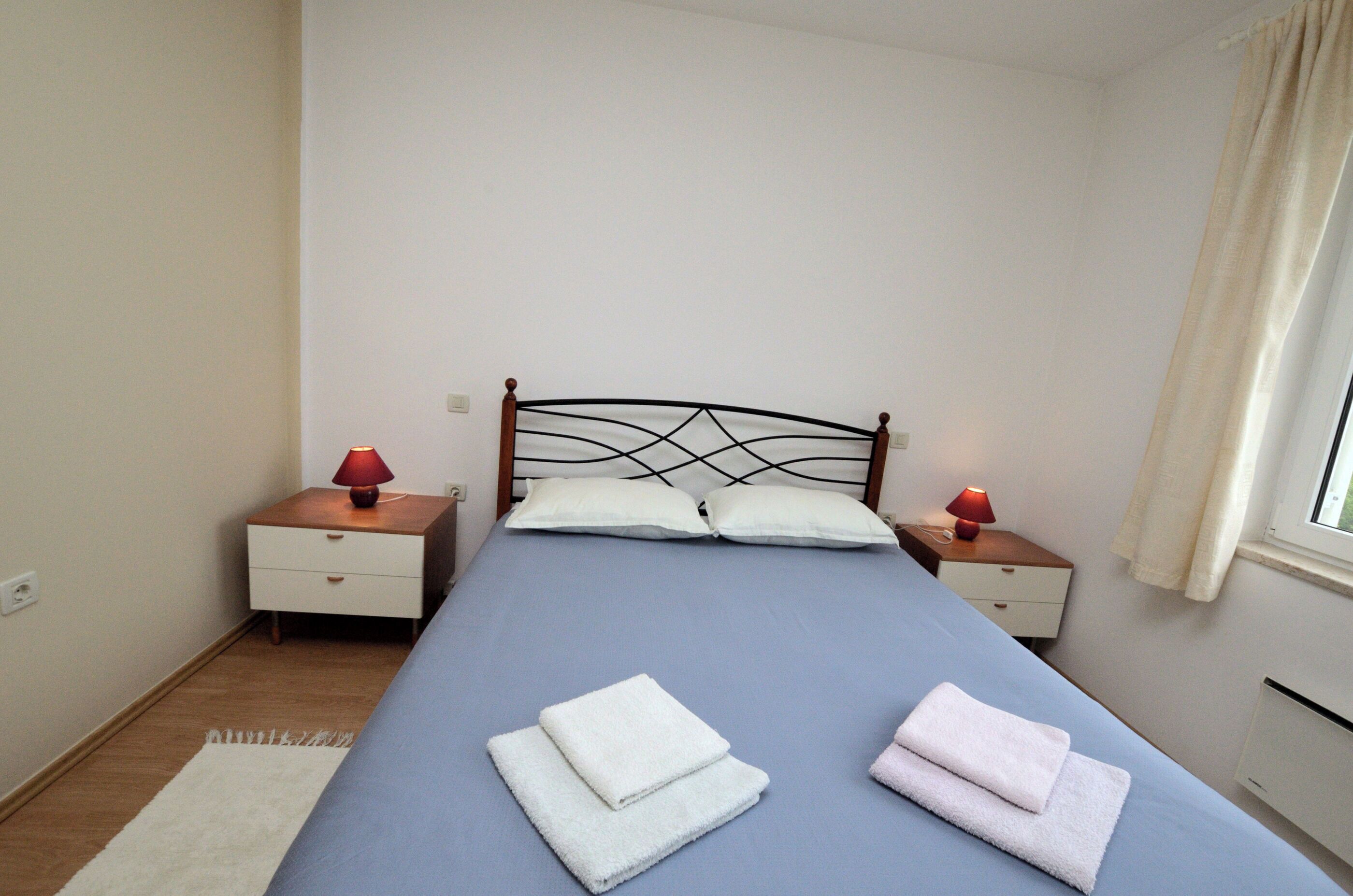 A neatly made bed with two pillows, two bedside tables with lamps, and a window with curtains.