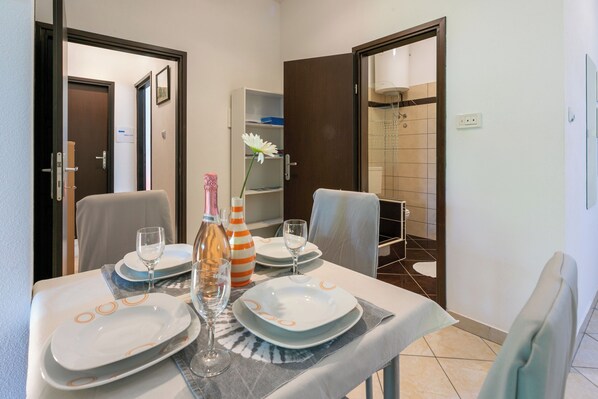 A dining area with a table set for four, including plates, glasses, and a bottle. There are chairs around the table, and a shower area is visible in the background. - Apartment Milojka (Crikvenica)