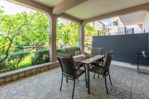 An outdoor patio with a stone floor, a table with chairs, and a view of a garden with trees and a fence. - Apartment Milojka (Crikvenica)