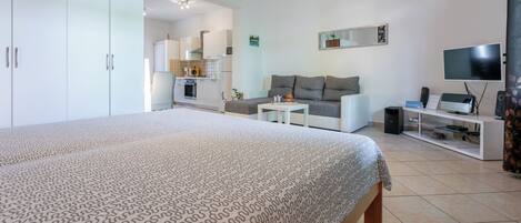 A modern bedroom with a large bed, a sofa, a TV, and a small kitchenette in the background.