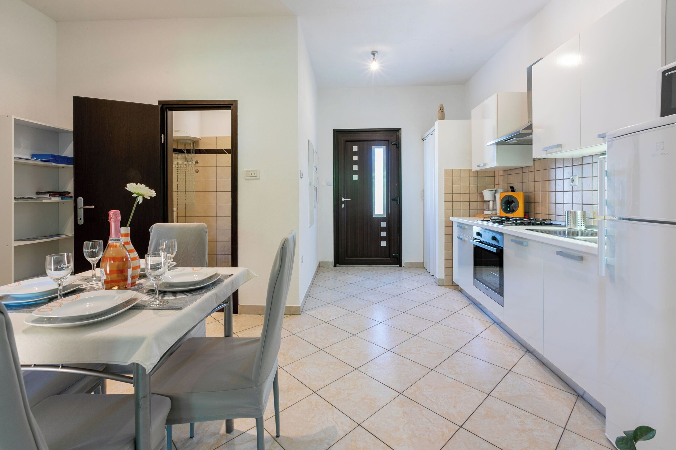 Une cuisine moderne avec des meubles blancs, une cuisinière et un réfrigérateur. On y trouve un coin repas avec une table dressée pour deux personnes, ainsi qu’une porte donnant sur une salle de bain.