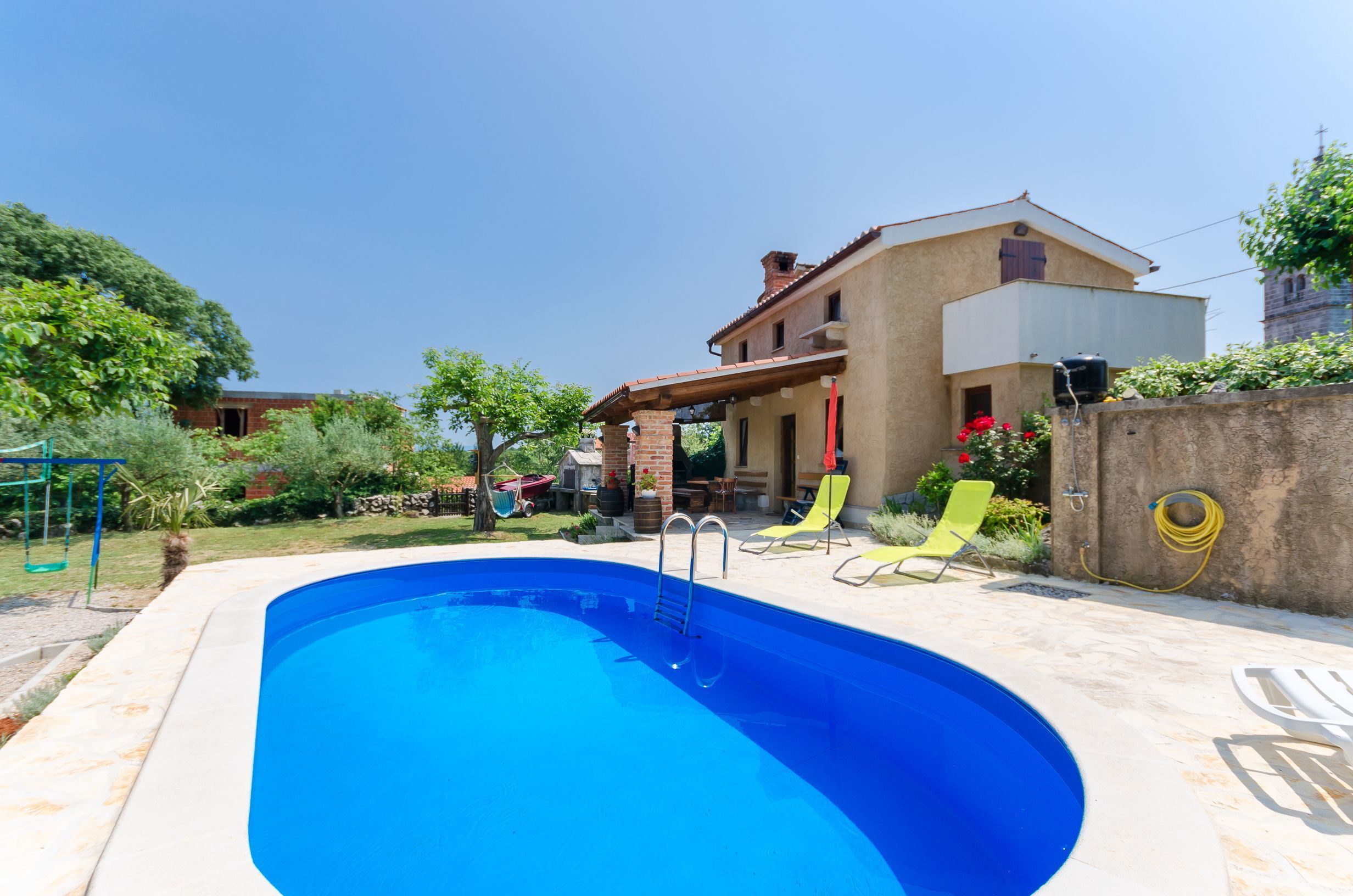 A swimming pool with a ladder, surrounded by a patio area with chairs and a house in the background.