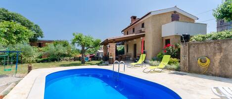 A swimming pool with a ladder, surrounded by a patio area with chairs and a house in the background.