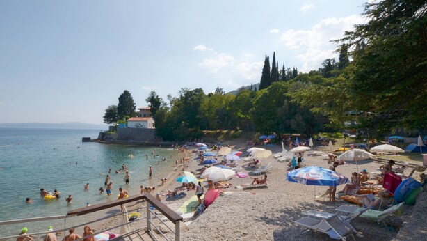 A beach with people swimming and sunbathing under umbrellas, a wooden deck, and a rocky shoreline. - Apartments Viktoria (Lovran)