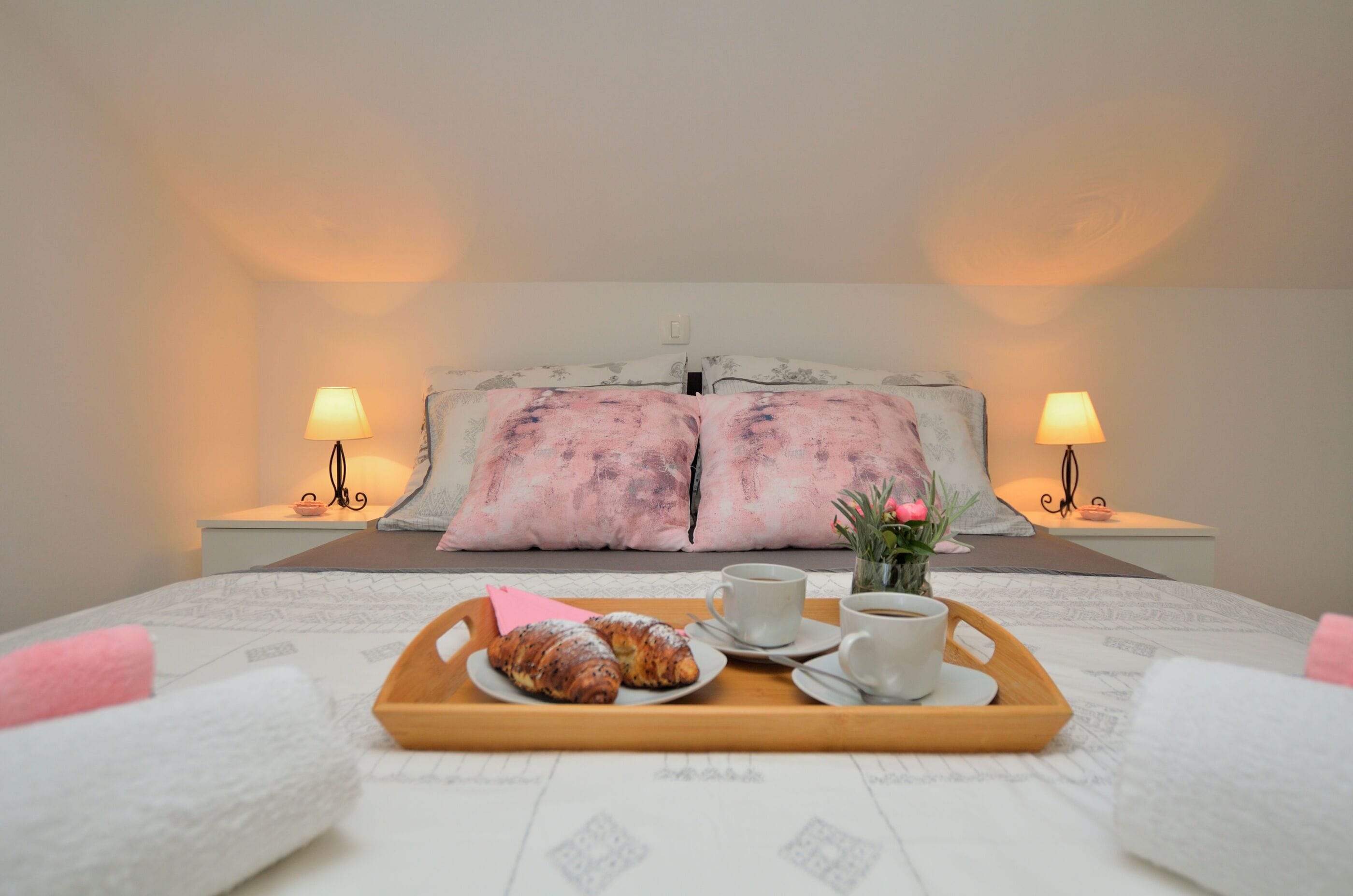 A neatly made bed with a tray of croissants and coffee cups, a vase with flowers, and two bedside lamps.
