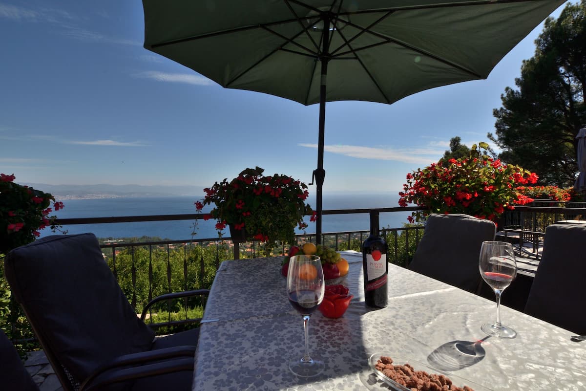 Uma mesa com uma garrafa de vinho, duas taças, uma tigela de frutas e uma rosa vermelha em um terraço com vista para o mar.