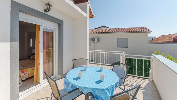A balcony with a table and chairs, a sliding glass door, and a view of a neighboring building. - Apartments Danica (Pirovac)