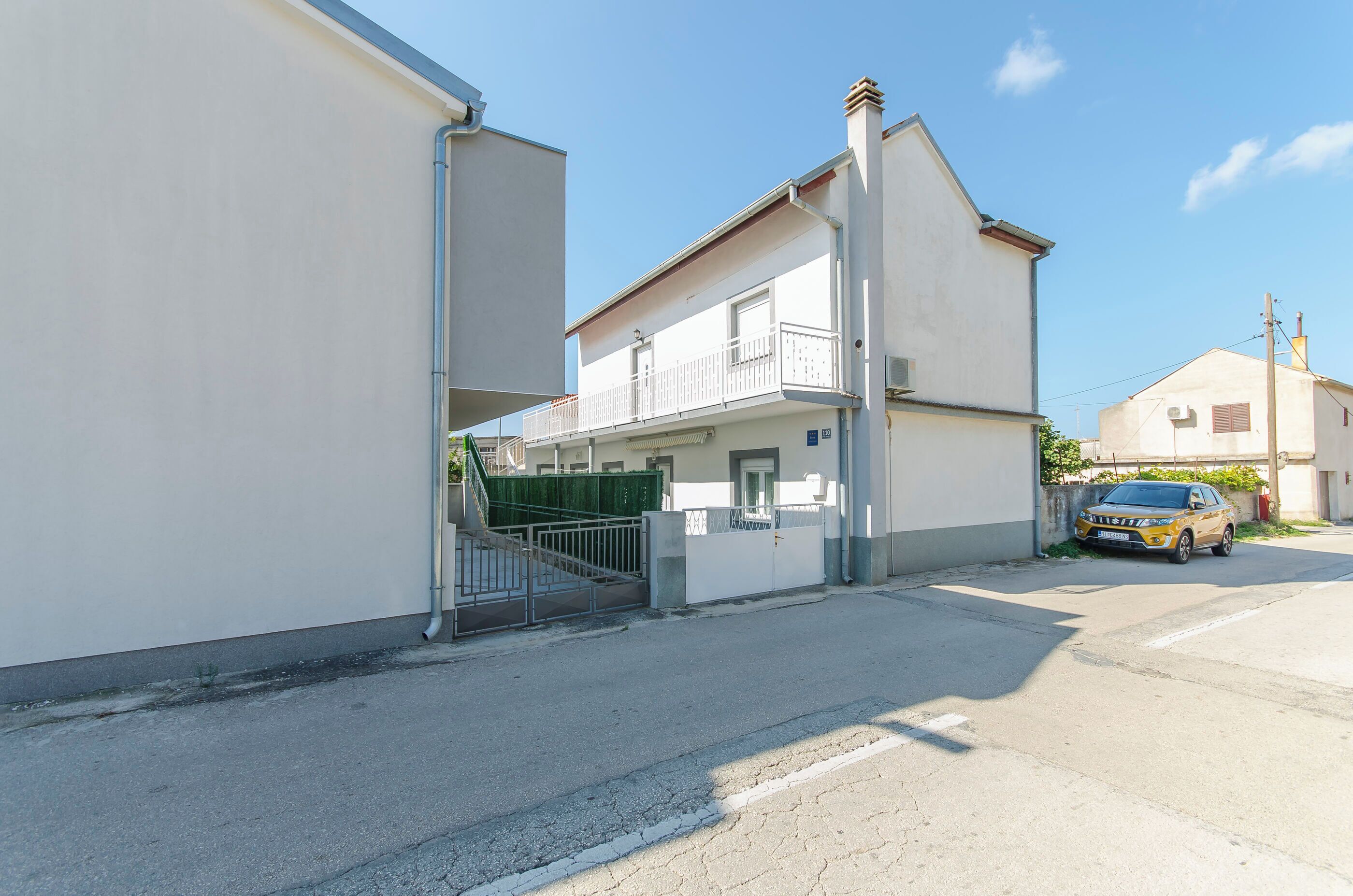 Eine Wohnstraße mit einem weißen Haus, das einen Balkon, ein grünes Tor und ein geparktes Auto zeigt.