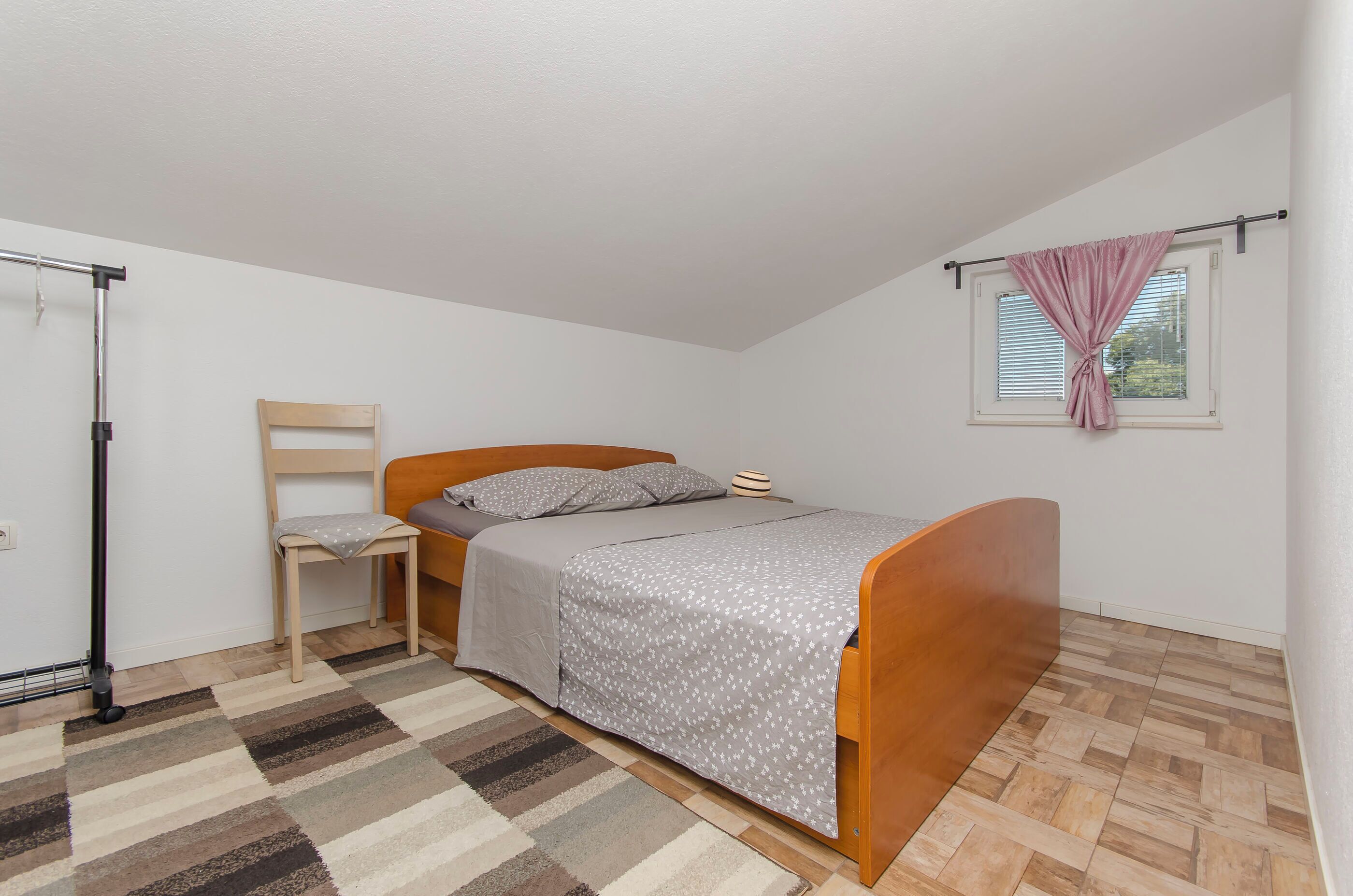 Een kleine, verzorgde slaapkamer met een enkel bed, een stoel, een raam met zonwering en een radiator.