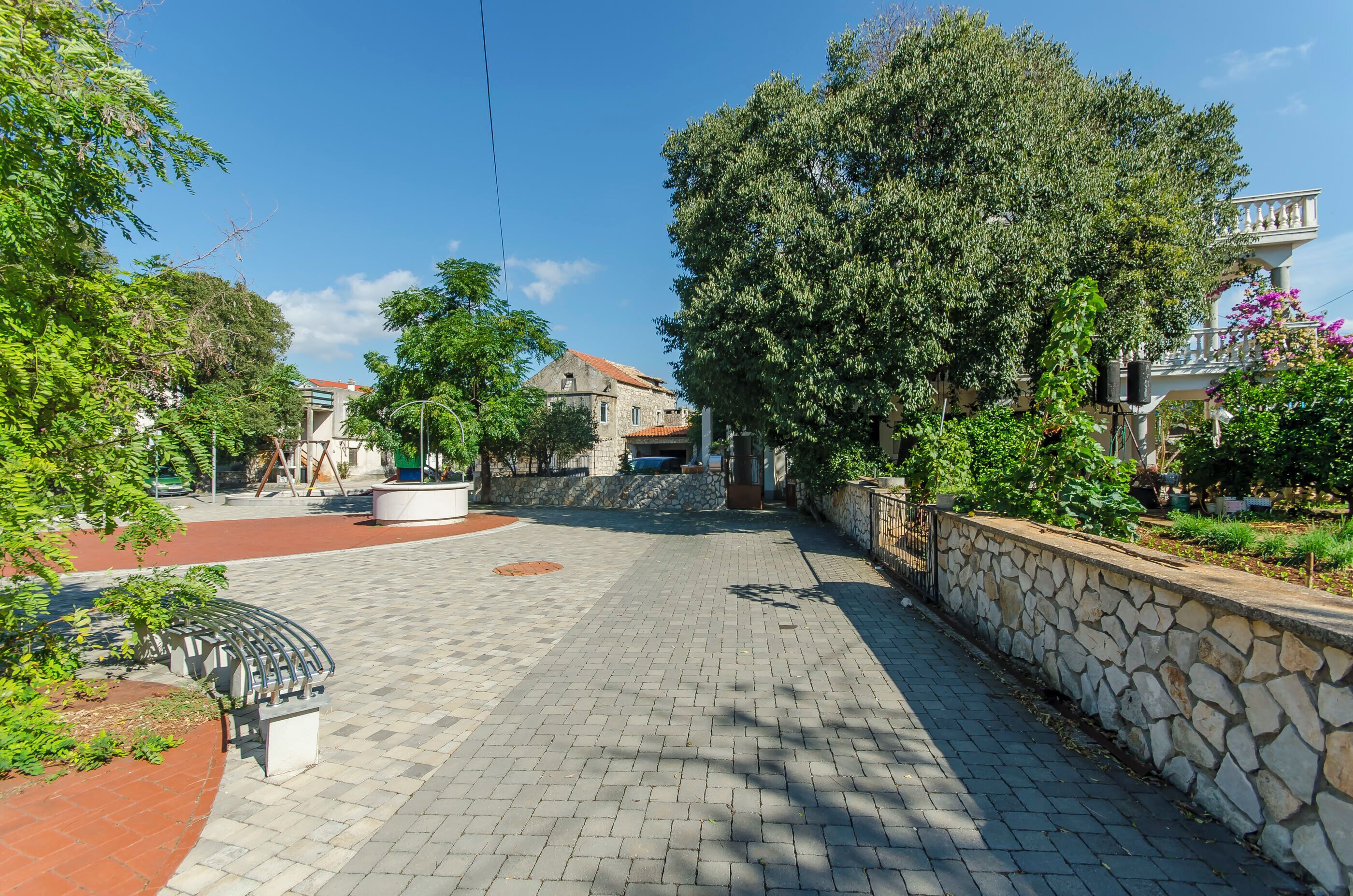 A paved walkway with stone walls and greenery, leading to a stone building with a red roof.