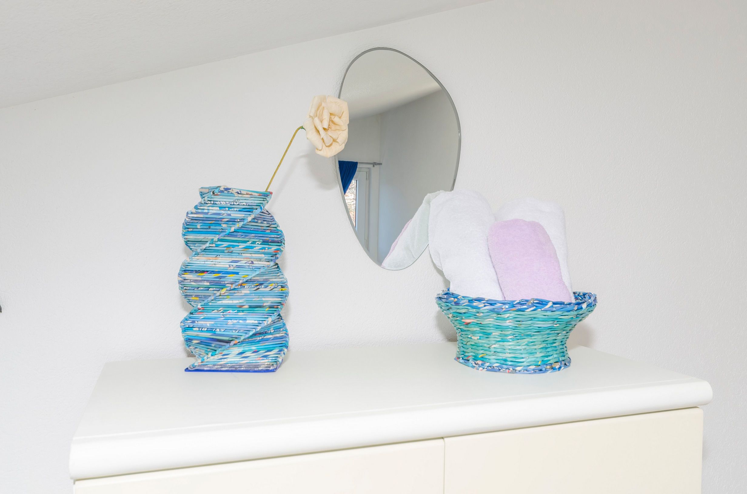 Ein Badezimmer-Waschtisch mit weißer Arbeitsplatte, eine blaue und weiße Vase mit einem einzelnen Blumenstiel und ein Korbwäscher mit einem weißen und einem rosafarbenen Handtuch.