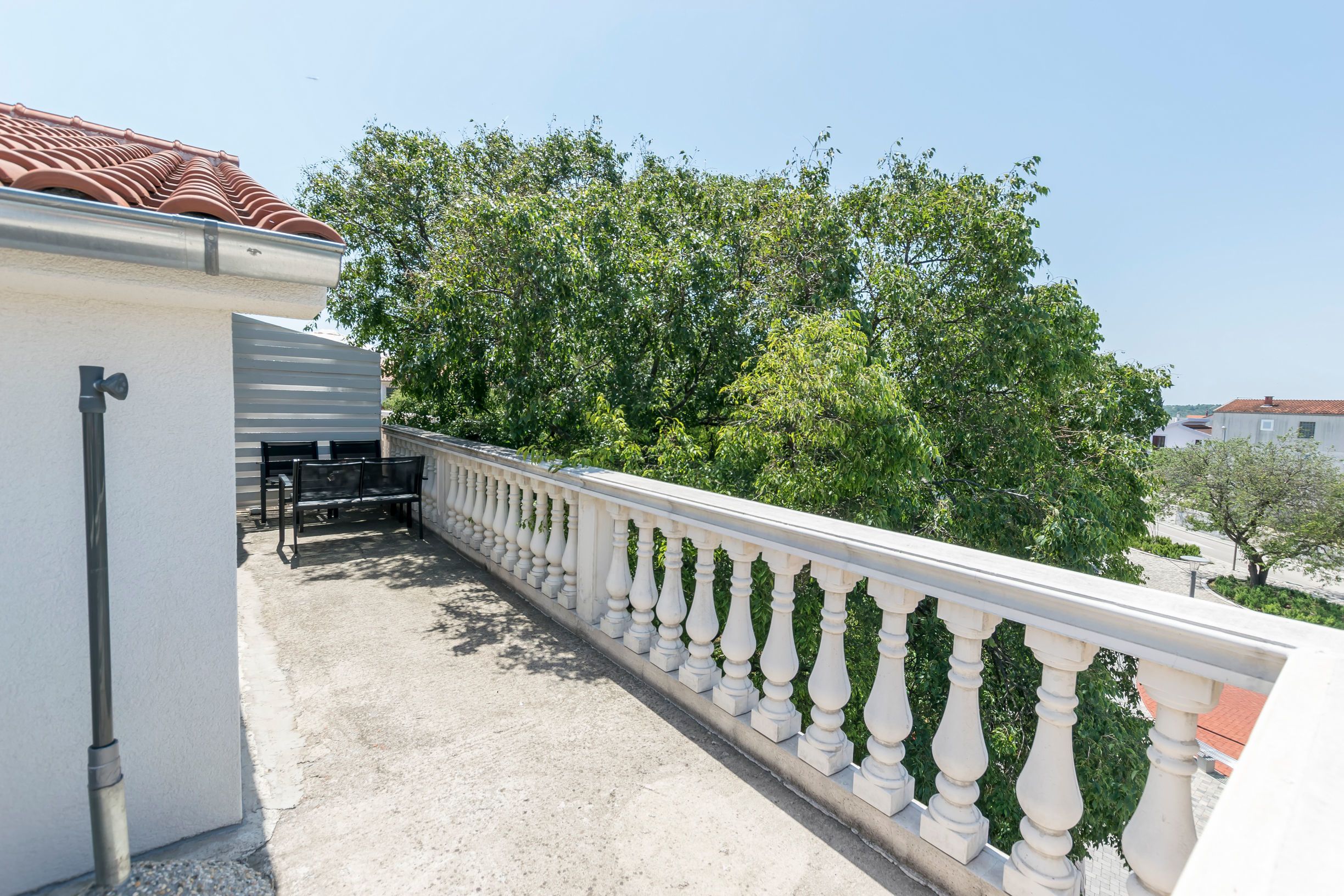 Un balcon doté d’une balustrade blanche, d’un sol en béton et offrant une vue sur les arbres et les bâtiments en arrière-plan.