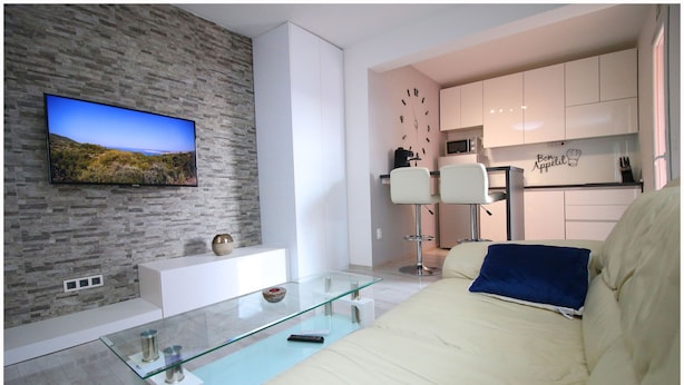 Una sala de estar moderna con un televisor de pantalla plana, una mesa de centro de vidrio, un sofá y una cocina con armarios blancos y un reloj.