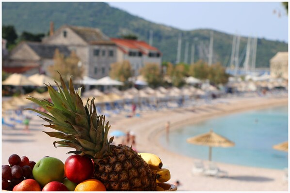 A beach with a pineapple, apples, and grapes in the foreground, and a building with a red roof in the background. - Apartments Eva (Primosten)