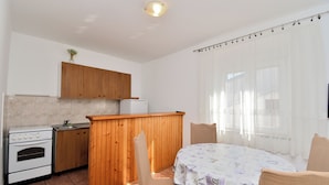 A compact kitchen with wooden cabinets, a white stove, and a small sink. There is a dining area with a table and chairs, and a window with sheer curtains.