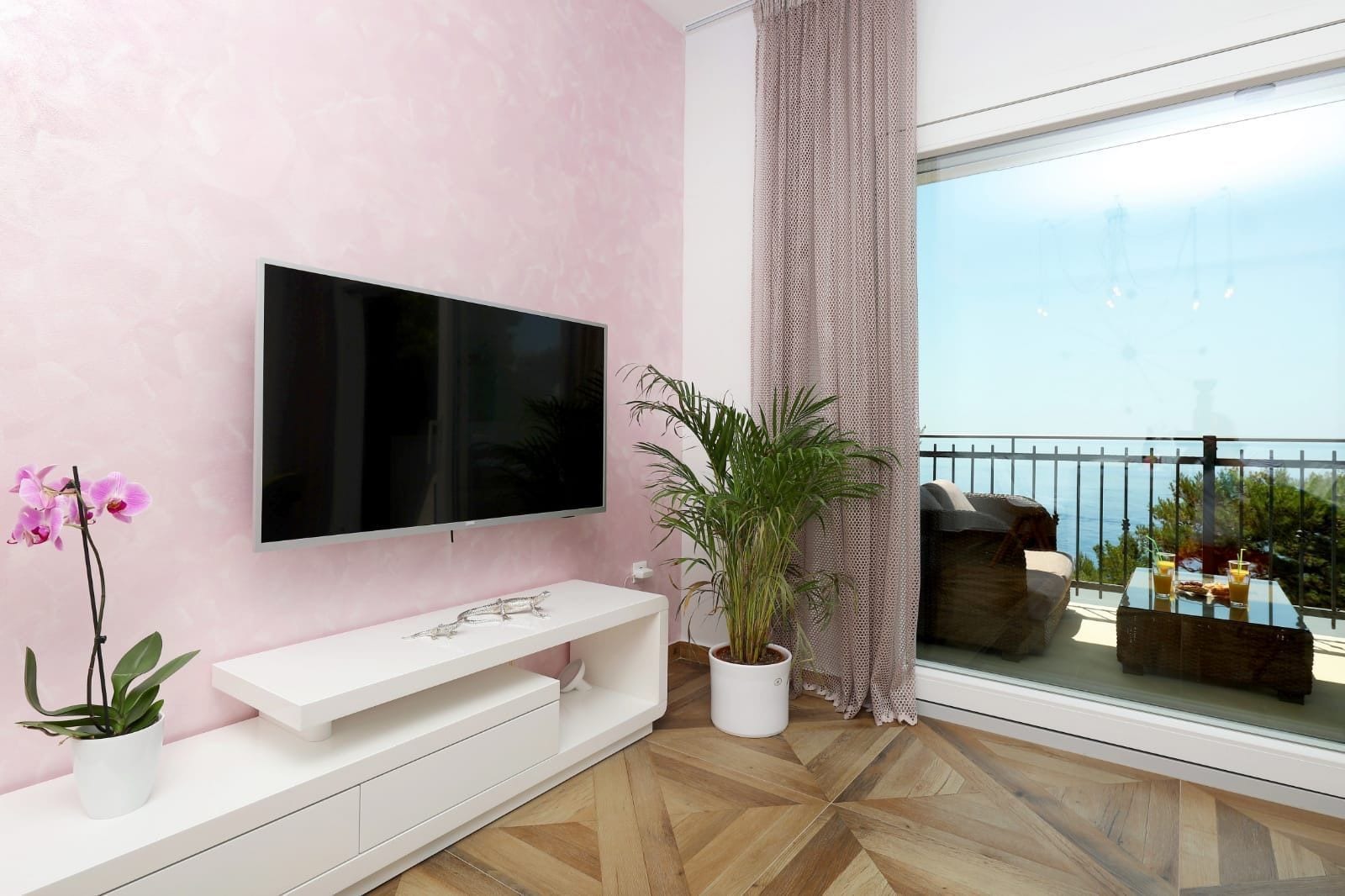 Un salon moderne doté d’un téléviseur à écran plat, d’un meuble de rangement blanc, de plantes en pot et d’un balcon avec des sièges d’extérieur.