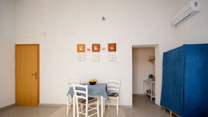 A room with a wooden door, a dining table with four white chairs, a blue tablecloth, and a fruit bowl. There are wall-mounted picture frames and an air conditioning unit. - Apartments Nina (Stinica)