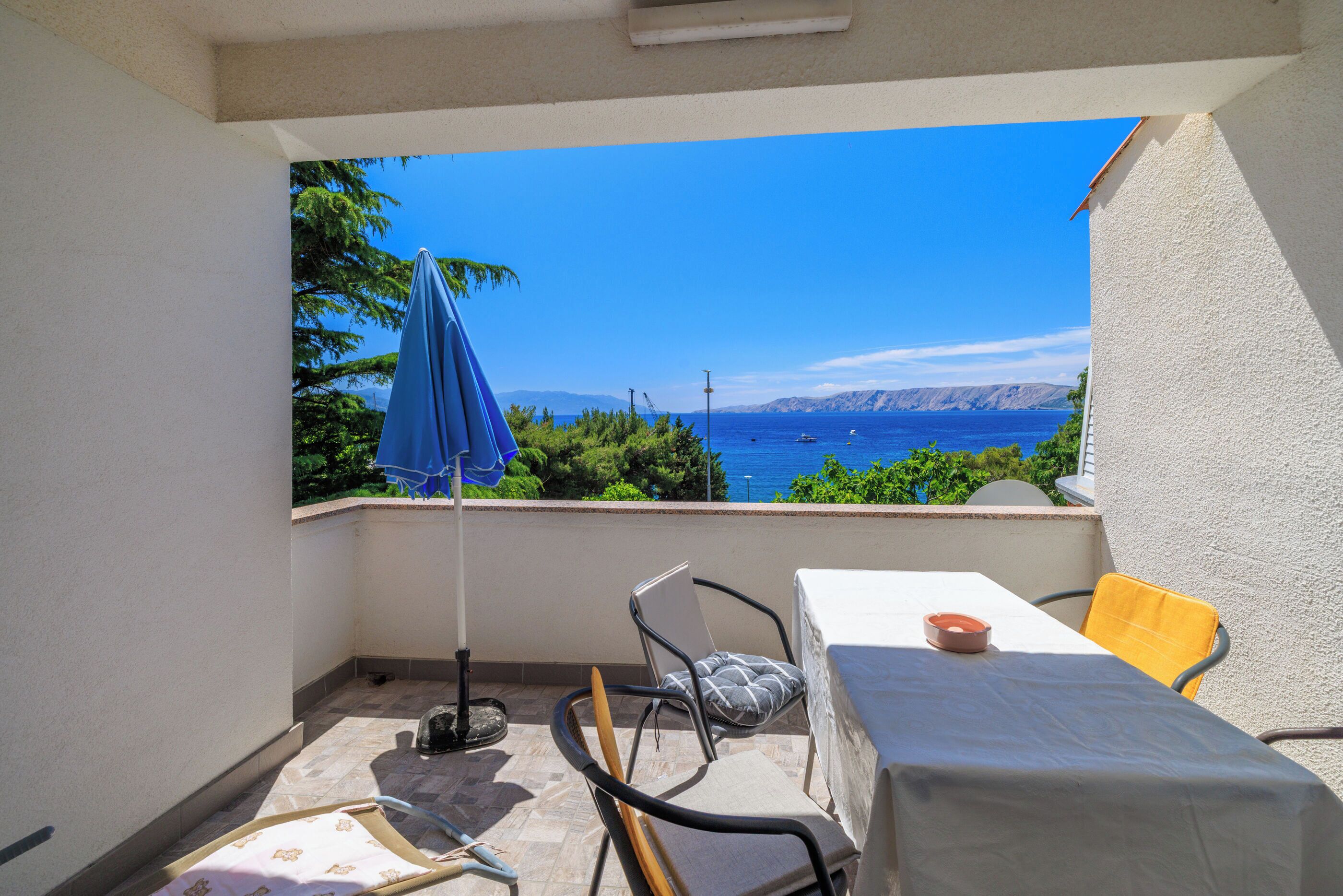 A balcony with a table, chairs, and an umbrella, overlooking a scenic view of the sea and mountains.
