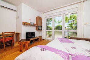 A bedroom with a bed, a desk, a television, and a window with a view of greenery.