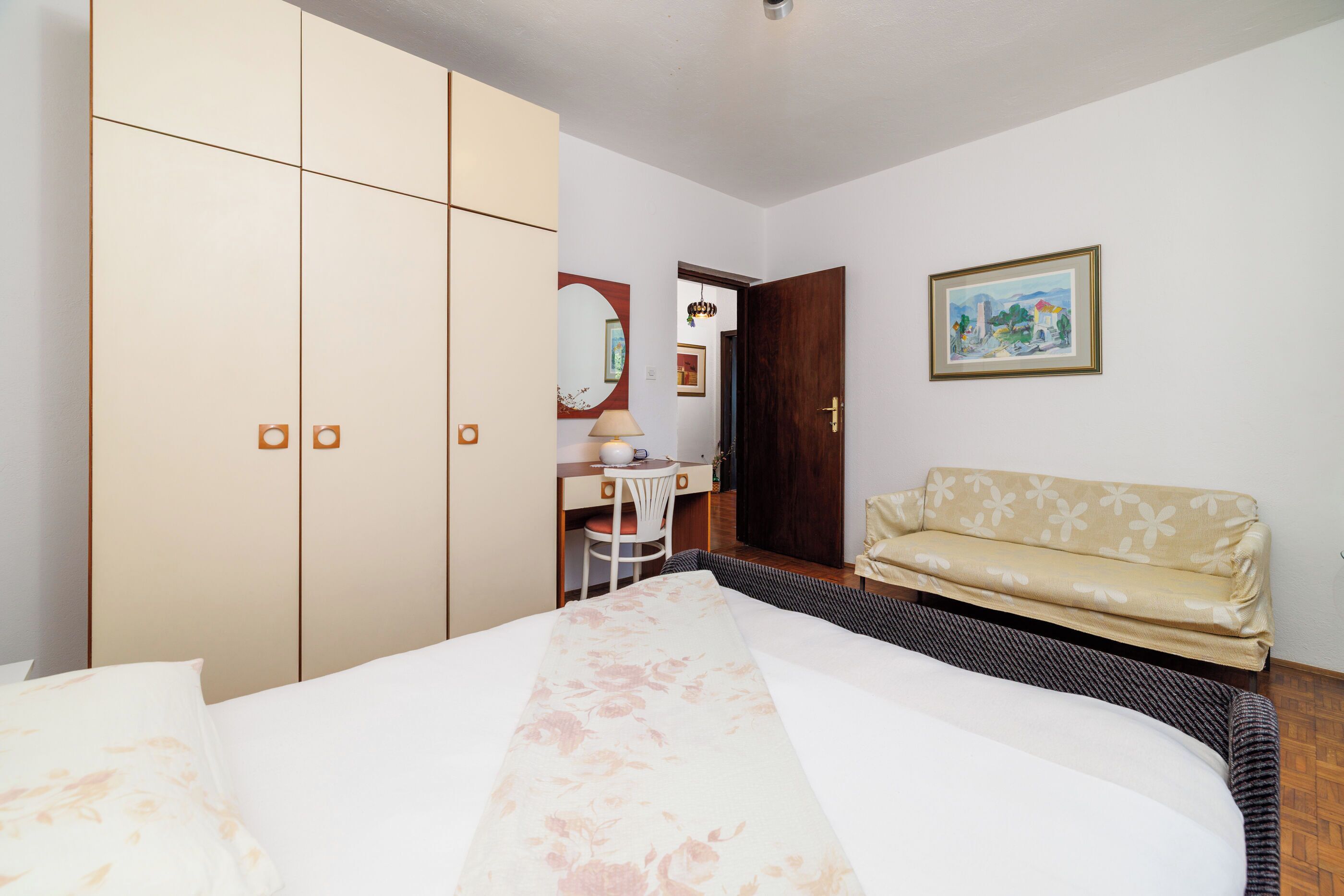 A bedroom with a bed, a wardrobe, a desk, a chair, a sofa, a mirror, and a framed picture on the wall.