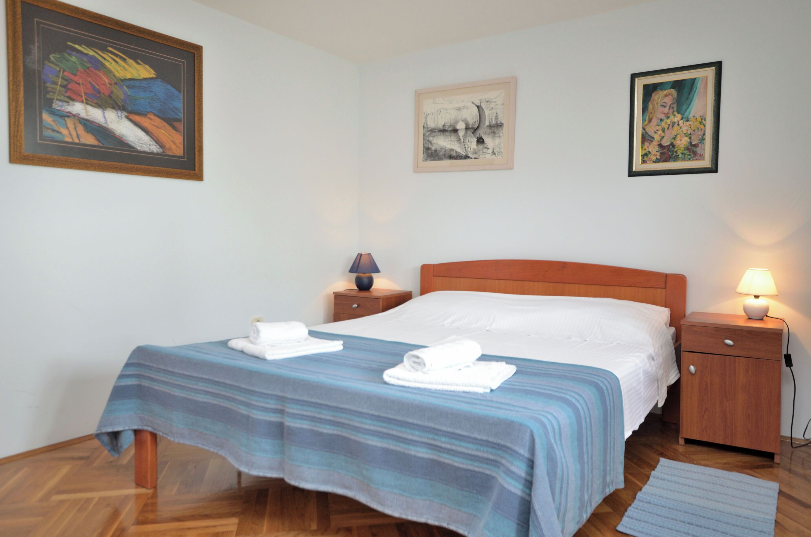 Un lit bien fait, recouvert d’une couverture à rayures, deux tables de chevet avec des lampes et trois œuvres d’art encadrées accrochées au mur.