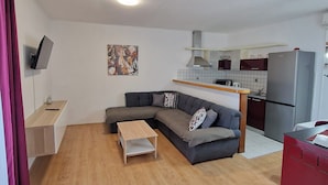 Un salon moderne avec un canapé d’angle gris, une table basse en bois et un coin cuisine équipé d’un réfrigérateur et d’un four à micro-ondes.