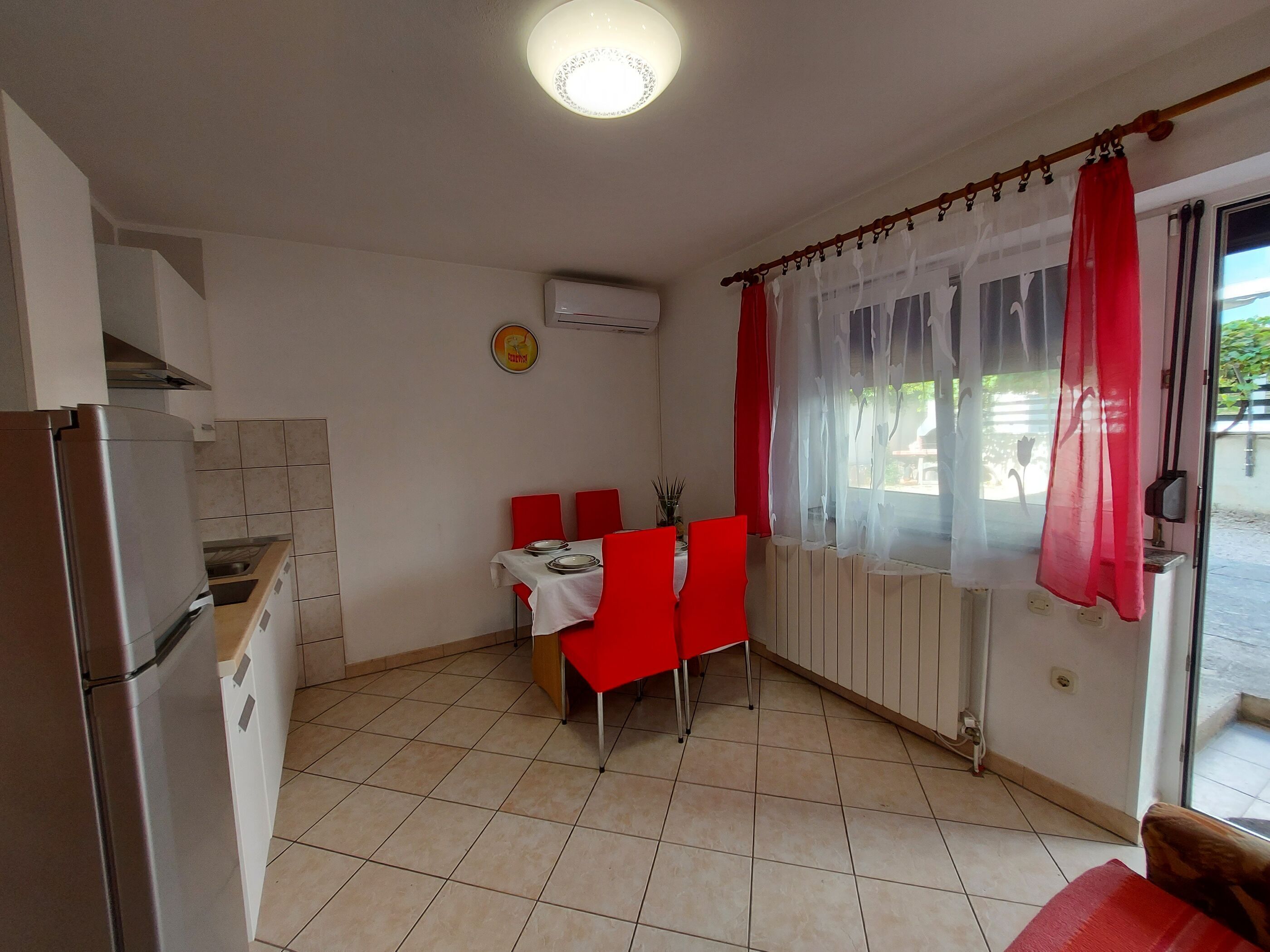 Eine kompakte Küche mit Kühlschrank, einem Esstisch mit roten Stühlen und einem Fenster mit transparenten Vorhängen.