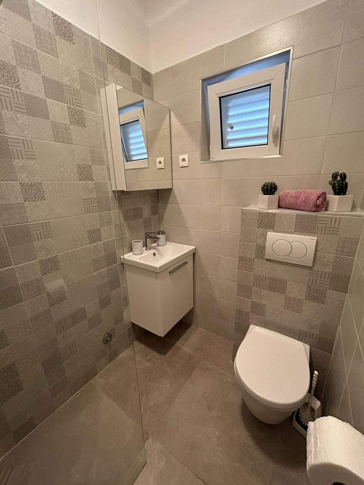 A small bathroom with a toilet, sink, and mirror. There are decorative tiles on the walls and a small window above the mirror.