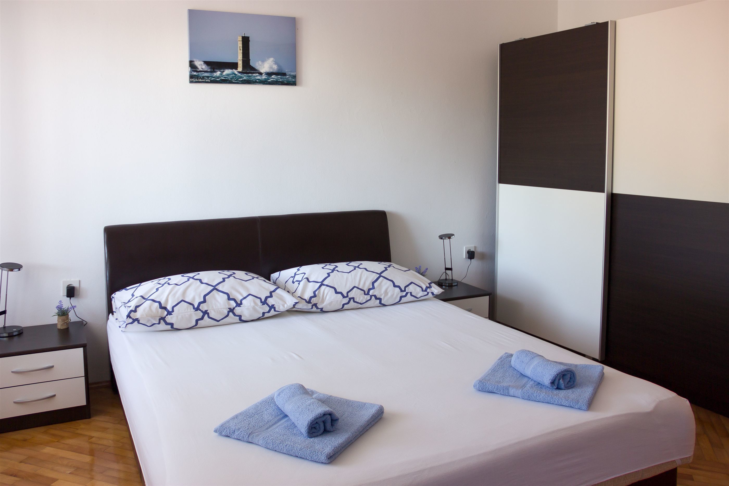 A neatly made bed with white sheets and blue towels, a dark headboard, and a wall-mounted picture of a lighthouse.