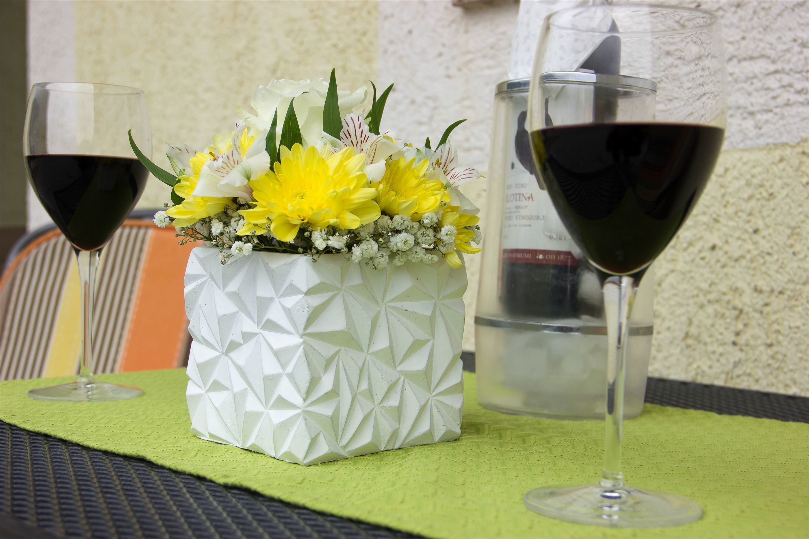 Una mesa dispuesta con dos copas de vino, un jarrón blanco con flores amarillas y un mantel individual verde.