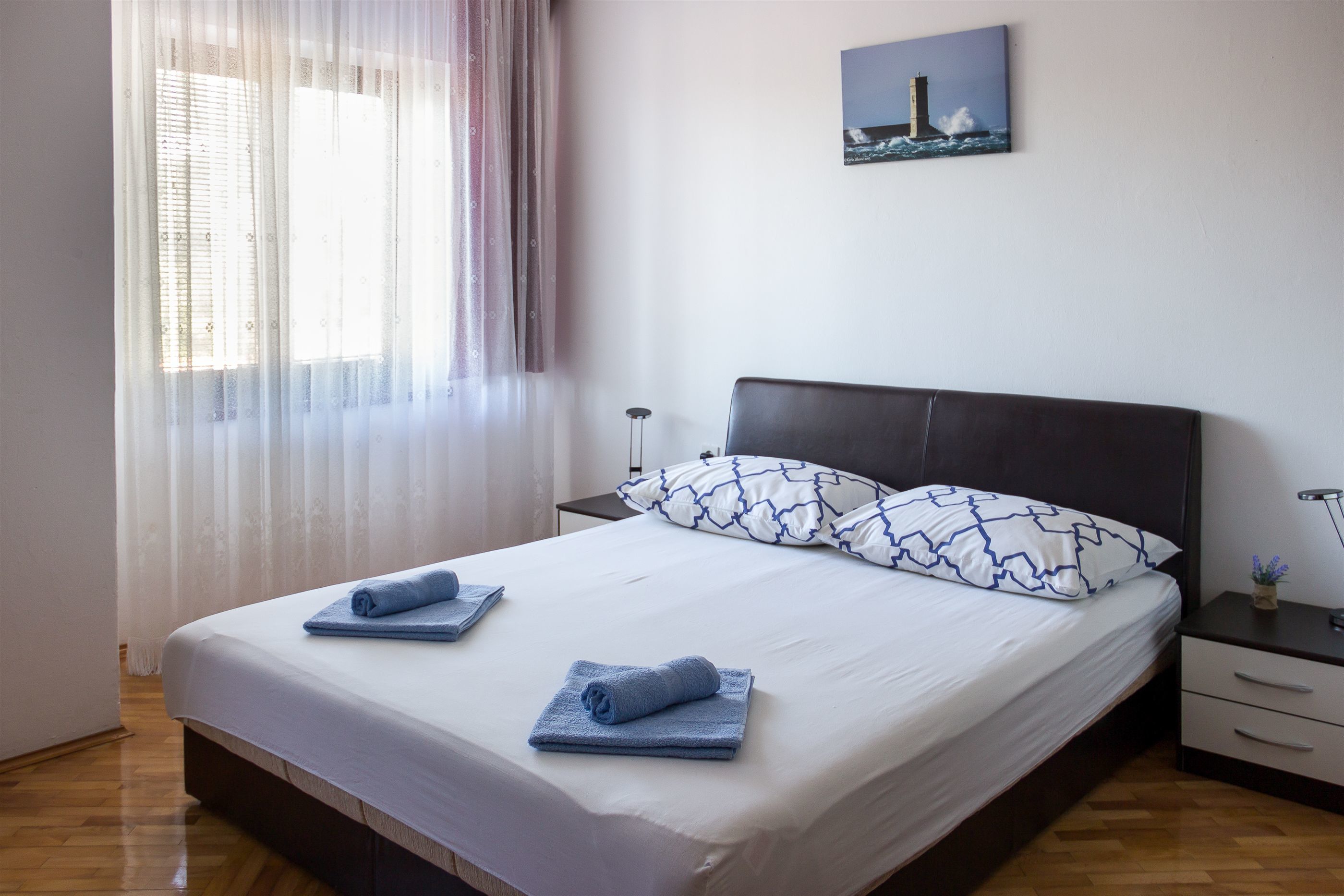 A neatly made bed with blue towels, a dark headboard, and a painting of a lighthouse on the wall.