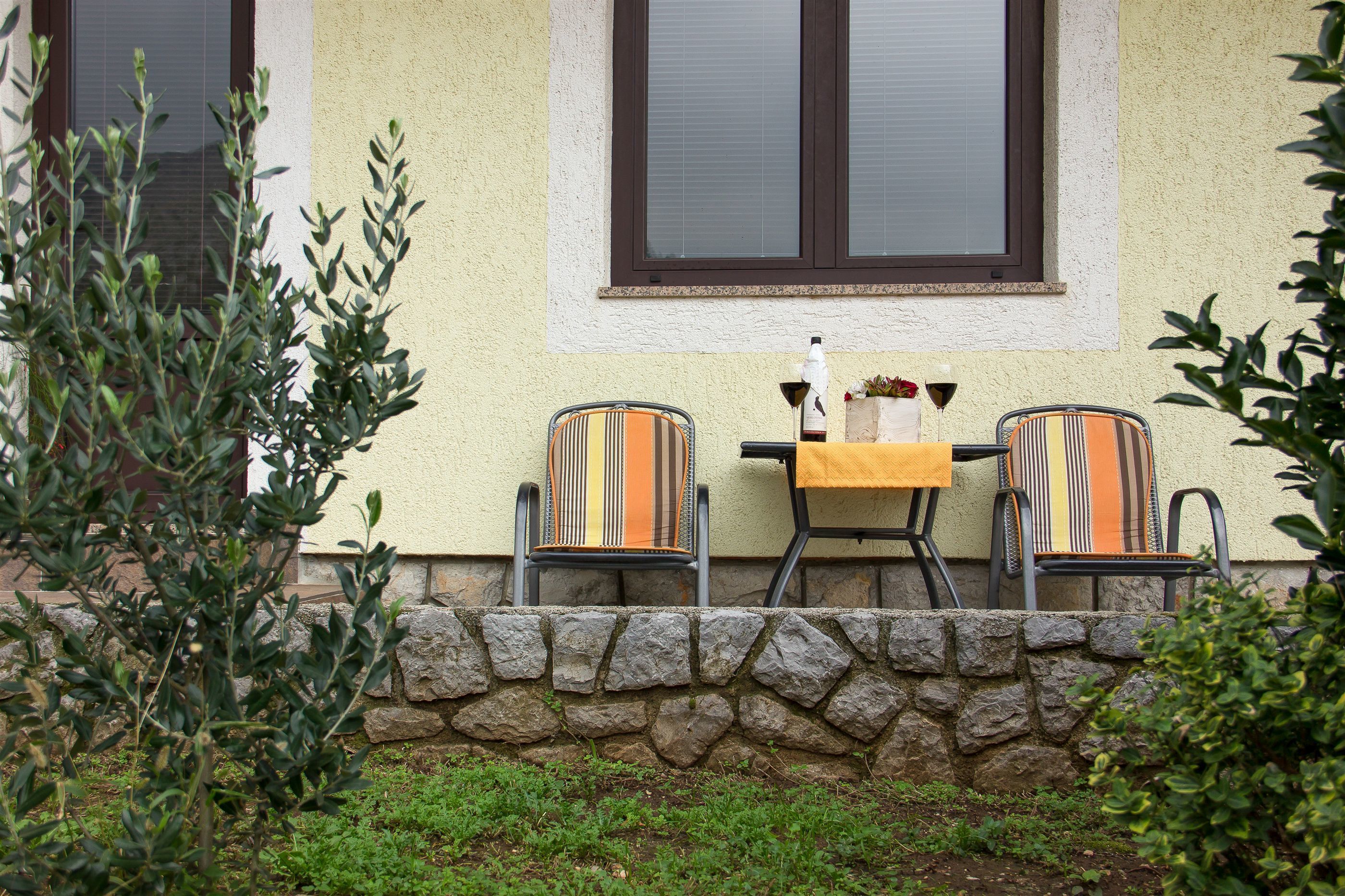 An outdoor seating area with two chairs and a table, a stone wall, and a window with blinds.
