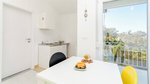 A modern kitchen with a white countertop and a sink, a dining table with a yellow chair, and a balcony with a view of trees. - Apartments Brulo (Razanj)