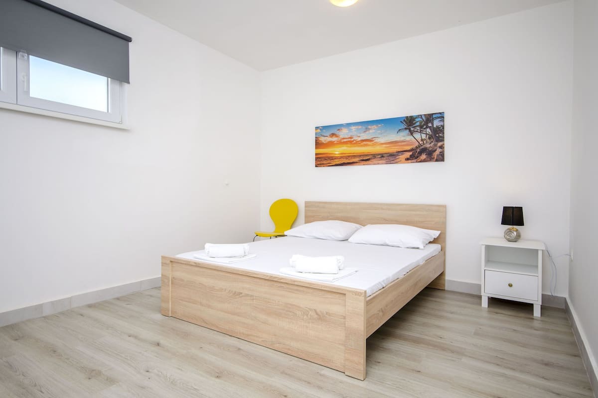 A bedroom with a wooden bed, white pillows, a nightstand with a lamp, and a painting of a sunset on the wall.