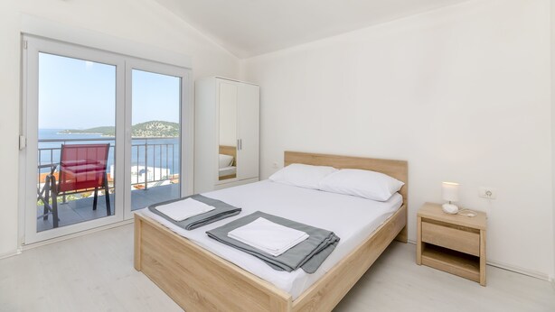A bedroom with a wooden bed, a bedside table with a lamp, and a red chair by a sliding glass door leading to a balcony with a view. - Apartments Brulo (Razanj)