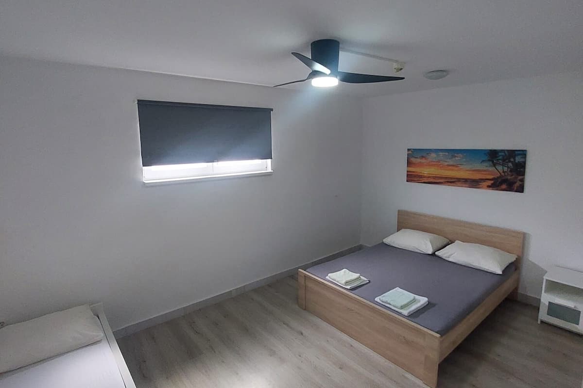 A bedroom with a bed, a bedside table, a ceiling fan, a window, and a painting on the wall.