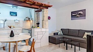 A modern living room with a grey sofa, a coffee table, and a TV. There's a dining area with a white table and chairs. The room has a stone fireplace and a ceiling light fixture. - Apartments Mila (Ubli)